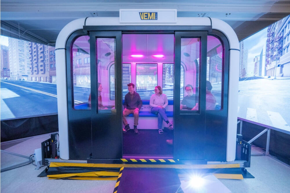 Two people sit inside a modern, enclosed vehicle simulator with sliding glass doors and purple lighting. The simulator is surrounded by large screens displaying a virtual cityscape, creating the illusion of being in an urban environment.
